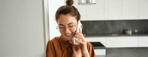 Portrait of lovely young woman answers phone call and smiling, leaning on wall, standing at home and talking on telephone photo