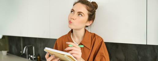 Portrait of cute young woman with notebook, sitting in the kitchen and writing her to do list, making tasks for week, puts notes in schedule or planner photo