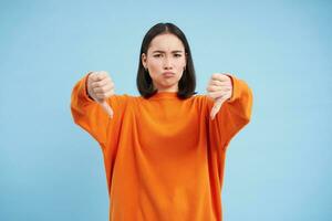 Very bad. Upset korean woman shows thumbs down, frowns and sulks disappointed, dislike smth, gives negative feedback, stands over blue background photo