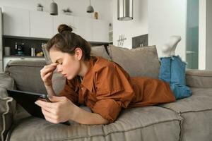 Portrait of woman resting on couch at home, rubs her eyes after reading too long on digital tablet, feeling tired, spending time in living room on sofa photo