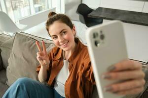 Positive and happy young woman taking selfie on smartphone, sitting on couch, showing peace, v-sign gesture photo