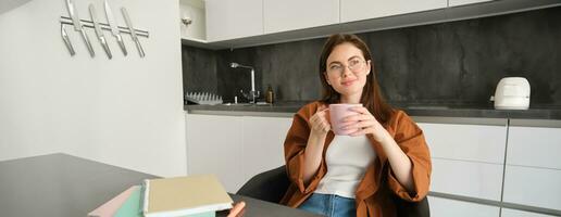 Portrait of smiling young woman in glasses, drinking coffee and looking outside window, sitting in kitchen with homework. Self-employed girl working from home, taking break, enjoying hot tea photo