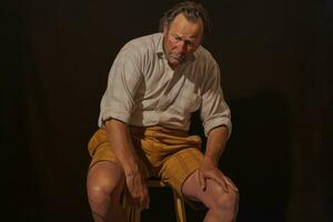 AI generated Portrait of an elderly man sitting on a chair on a dark background, a man holds on to sore knee, AI Generated photo