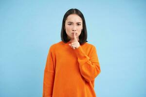 Angry young woman say shh, shows finger on lips and frowns in disapproval, tells to be quiet, hush gesture, shushing at someone, blue background photo