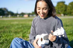 retrato de asiático niña alumno, jugando ukelele y canto en parque, sentado solo en cobija y disfrutando haciendo música foto