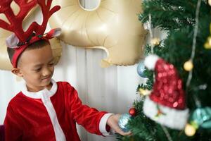 Merry Christmas 2024 concept Asian boy decorates Christmas tree with colorful Christmas balls. photo