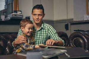 joven padre y su elegante pequeño hijo en el barbería en el esperando habitación. foto