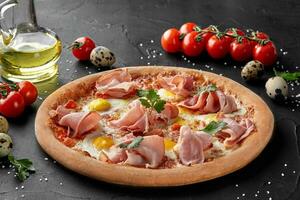 Pizza with quail eggs and ham on black surface photo