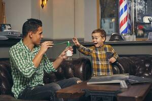 joven padre y su elegante pequeño hijo en el barbería en el esperando habitación. foto