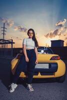 Female in sunglasses, white top, blue jeans and sneakers is posing outdoors near yellow car with two glass bottles of soda on its trunk. Mock up photo