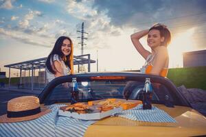 amigos hembras son sonriente, posando en amarillo coche cabrio con francés papas fritas, Pizza y soda en vaso botellas en sus trompa. rápido alimento. Copiar espacio foto