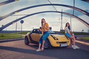 Gorgeous models in casual outfit are enjoying drinks in paper cups and hamburgers while sitting on the hood of yellow car roadster. Fast food photo