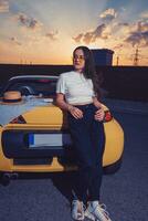 Young lady in sunglasses, white top, blue jeans and sneakers is posing outdoors near yellow car roadster with pizza on its trunk. Fast food photo