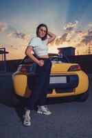 atractivo niña en Gafas de sol, blanco arriba, negro pantalones y zapatillas es posando al aire libre cerca amarillo coche cabriolé con dos botellas de soda en sus maletero foto