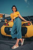 Young model in blue jeans, orange top and sunglasses. She is holding paper cup of coffee, leaning on yellow car at parking lot. Full length, mock up photo