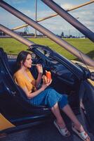 Woman in blue jeans and orange top is eating french fries from paper package and holding hamburger while sitting in yellow car cabrio Copy space photo