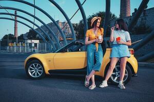 joven señoras son sonriente, disfrutando francés papas fritas desde papel paquete y bebidas en papel tazas, posando cerca amarillo coche. rápido alimento. Copiar espacio foto