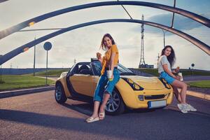 Ladies are smiling, holding hamburgers and beverages in paper cups while sitting on the hood of yellow car cabrio. Fast food. Copy space photo