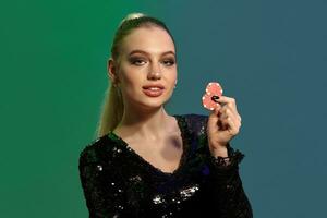 Blonde girl in jewelry and black sequin dress. She smiling, showing two red chips, posing on colorful background. Gambling, poker, casino. Close-up photo