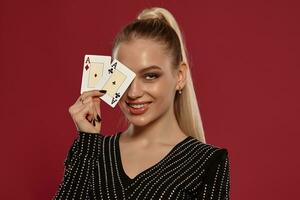 Blonde lady in black dress in rhinestones. She is smiling, showing two aces, posing against red background. Gambling, poker, casino. Close-up photo