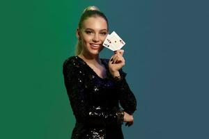 Blonde woman in jewelry and black sequin dress. She smiling, showing two playing cards, posing on colorful background. Poker, casino. Close-up photo