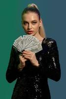 Blonde woman in jewelry and black sequin dress. She is holding fan of hundred dollar bills, posing on colorful background. Poker, casino. Close-up photo