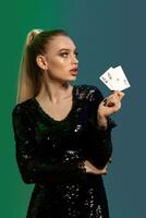 Blonde model in jewelry and black sequin dress. She is showing two playing cards, posing on colorful background. Gambling, poker, casino. Close-up photo
