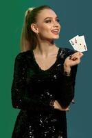 Blonde lady in jewelry and black sequin dress. She smiling, showing two playing cards, posing on colorful background. Poker, casino. Close-up photo