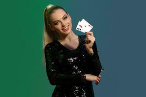 Blonde female in jewelry and black sequin dress. She smiling, showing two playing cards, posing on colorful background. Poker, casino. Close-up photo