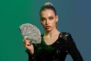 Blonde woman in jewelry and black sequin dress. She is showing fan of hundred dollar bills, posing on colorful background. Poker, casino. Close-up photo