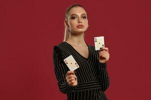 rubia niña en negro vestir en pedrería. demostración dos jugando tarjetas, posando en contra rojo antecedentes. juego entretenimiento, póker, casino. de cerca foto