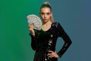 Blonde woman in jewelry and black sequin dress. She is showing fan of hundred dollar bills, posing on colorful background. Poker, casino. Close-up photo