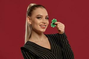 Blonde model with ponytail, in black dress in rhinestones. She smiling, showing two green chips, posing on red background. Poker, casino. Close-up photo