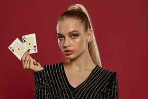 Blonde model in black dress in rhinestones. Showing two playing cards, posing against red background. Gambling entertainment, poker, casino. Close-up photo