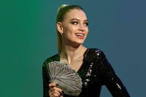 Blonde female in jewelry and black sequin dress. She is smiling, showing some cash while posing against colorful background. Poker, casino. Close-up photo