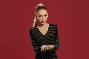 Blonde girl with ponytail, in jewelry and black dress. She is showing handful of colorful chips, posing on red background. Poker, casino. Close-up photo