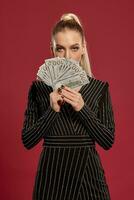 Blonde lady with ponytail, in black dress in rhinestones. Showing fan of hundred dollar bills, posing on red background. Poker, casino. Close-up photo