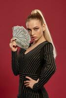 Blonde female with ponytail, in black dress in rhinestones. She is holding some money, posing sideways on red background. Poker, casino. Close-up photo