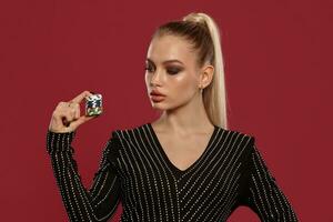 Blonde female with ponytail, in jewelry and black dress in rhinestones. She showing stack of chips, posing on red background. Poker, casino. Close-up photo