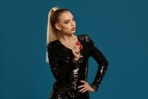 Blonde girl in black sequin dress showing two red chips, posing against blue background. Gambling entertainment, poker, casino. Close-up photo