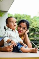 amoroso mamá que lleva de su bebé a hogar balcón. brillante retrato de contento mamá participación niño en su manos. madre con su pequeño 1 año antiguo hijo. foto