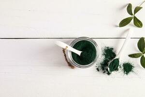 Green powder spirulina algae in glass jars and spoon on white wooden background. Top view. A copy space. high content of multivitamins photo