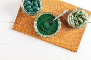 Green spirulina powder, round tablets, capsules in various glass bowls and jars on a wooden stand. Top view. A super food supplement. photo