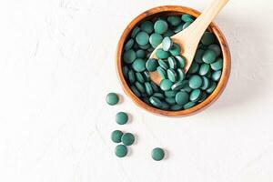 Round green spirulina algae pills in wooden bowl and spoon on white background with copy space. Top view. super product. photo