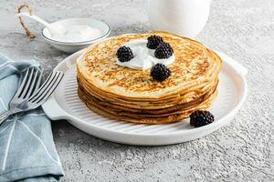 A large stack of delicious hot pancakes with sour cream and blackberries on a beautiful white serving tray with cutlery and a plot. Front view. photo