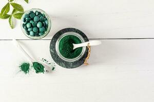 Green powder and spirulina algae tablets in glass jars and spoon on white wooden background. Top view. A copy space. high content of multivitamins. photo