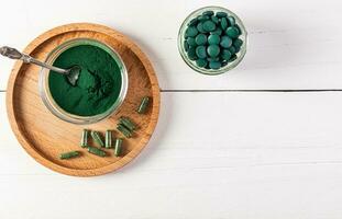 Natural Organic Green Spirulina Algae Powder In Glass Jar, Round Tablets, Capsules On Wooden Round Tray And White Background. Top view. photo