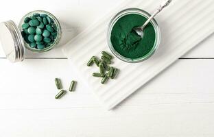 Spirulina powder, tablets, capsules in various jars on white wooden background and ceramic stand. Top view. Healthy nutritional supplements . photo