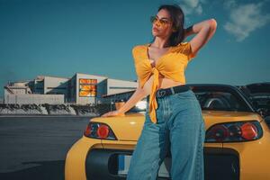 Woman in blue jeans, orange top and sunglasses is posing near yellow car cabriolet with paper cup of coffee on trunk at parking lot. Copy space photo