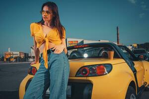 Lady in blue jeans, orange top and sunglasses is posing leaning on yellow car roadster with paper cup of coffee on trunk at parking lot. Close up photo